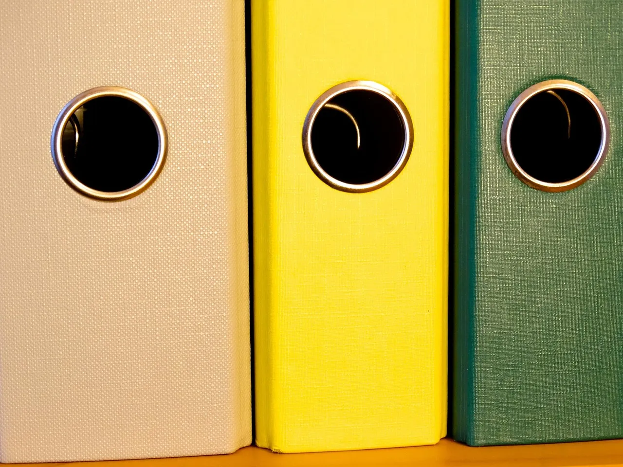 A close view of the spines of three unlabelled lever arch files standing side by side, cream, yellow and green, each with a round metal-rimmed finger hole.