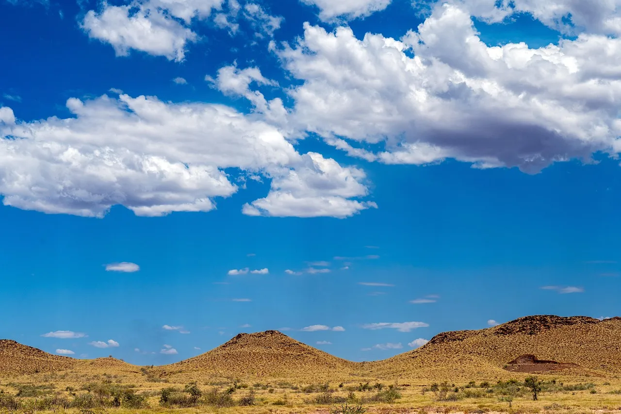 Low red-brown hills under a deep blue sky with white clouds, with dry scrub in the foreground.