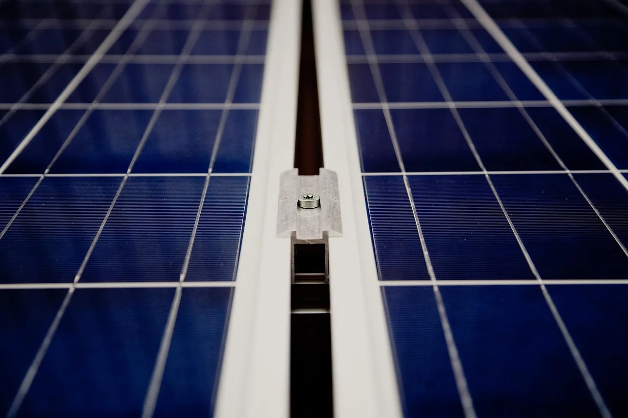 Close view of two dark blue solar panels, their cells ruled in a silver grid, held to a mounting rail between them by a small metal clamp and screw