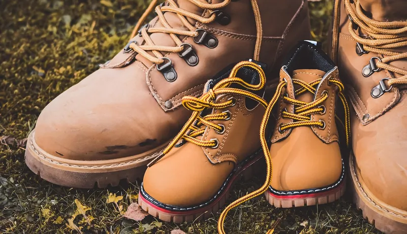 A small pair of tan lace-up children’s boots with yellow laces, standing on grass between two large worn adult boots