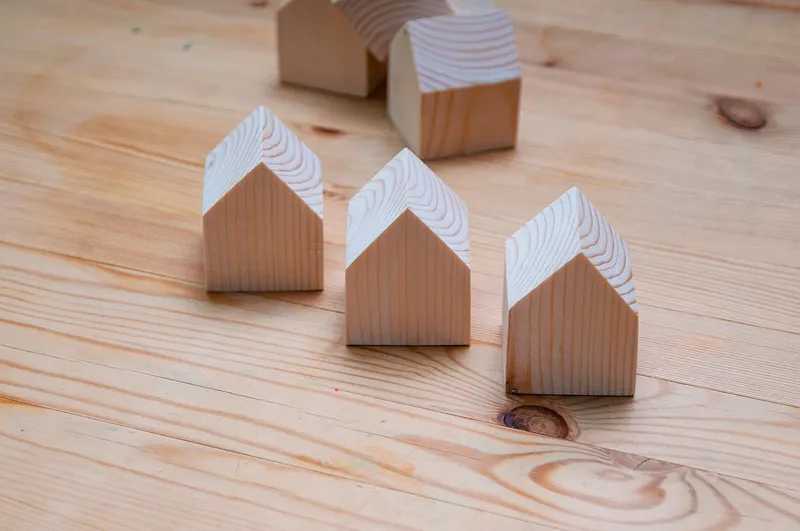 Five small unpainted wooden house blocks with pitched roofs standing on a pale pine tabletop