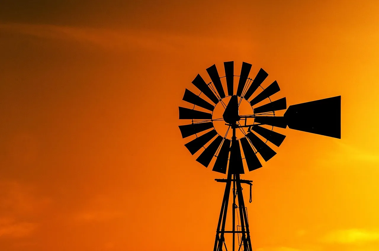 The silhouette of a farm windpump, its bladed wheel and tail vane black against an orange sky.