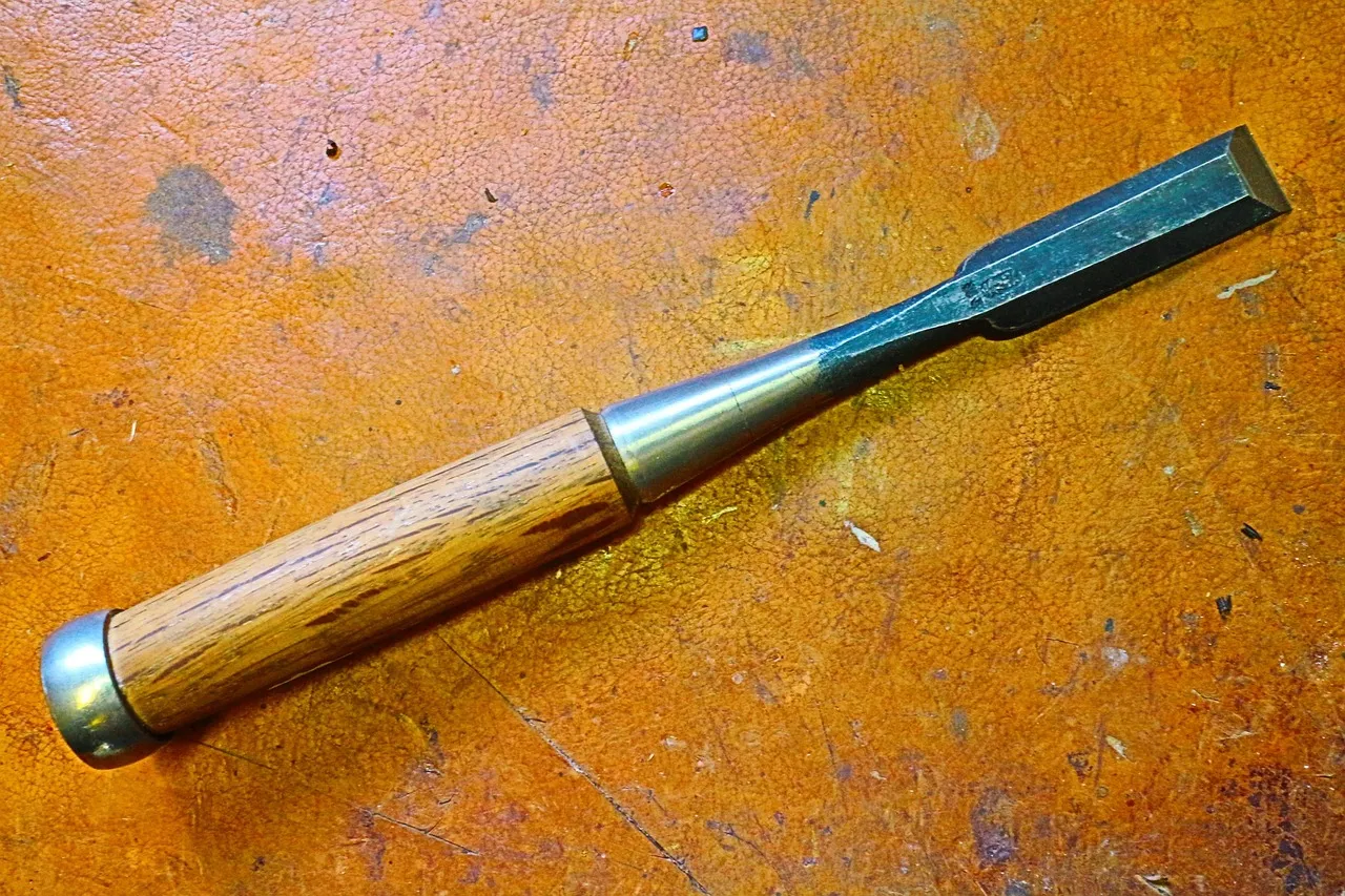 A wooden-handled chisel with a steel ferrule lying on a worn orange bench top