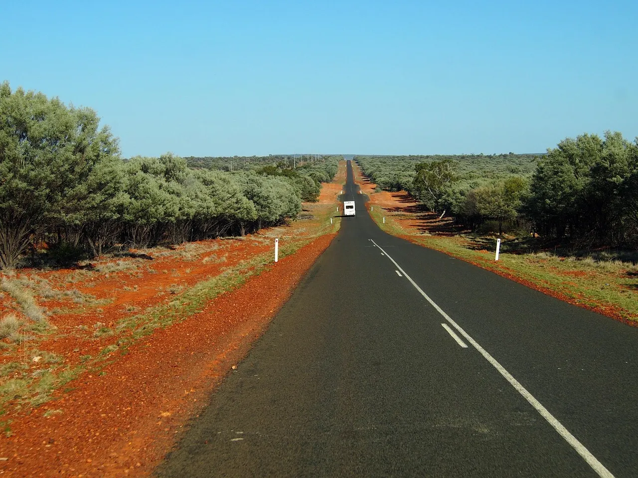 A straight sealed road running between grey-green scrub and red earth to the horizon, with a single caravan far ahead.