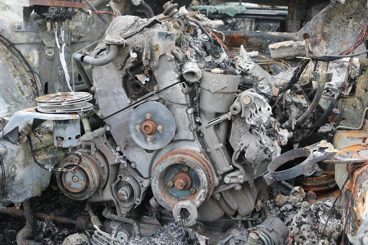 A burnt-out car engine and its pulleys, grey with ash, surrounded by melted wiring and scorched metal.
