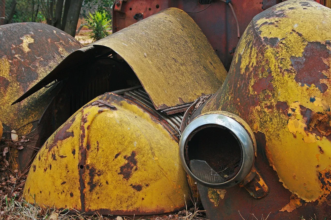 The front of an old vehicle rusting in long grass: rounded yellow mudguards flaking to bare rust, a buckled bonnet panel and an empty chrome headlamp ring.