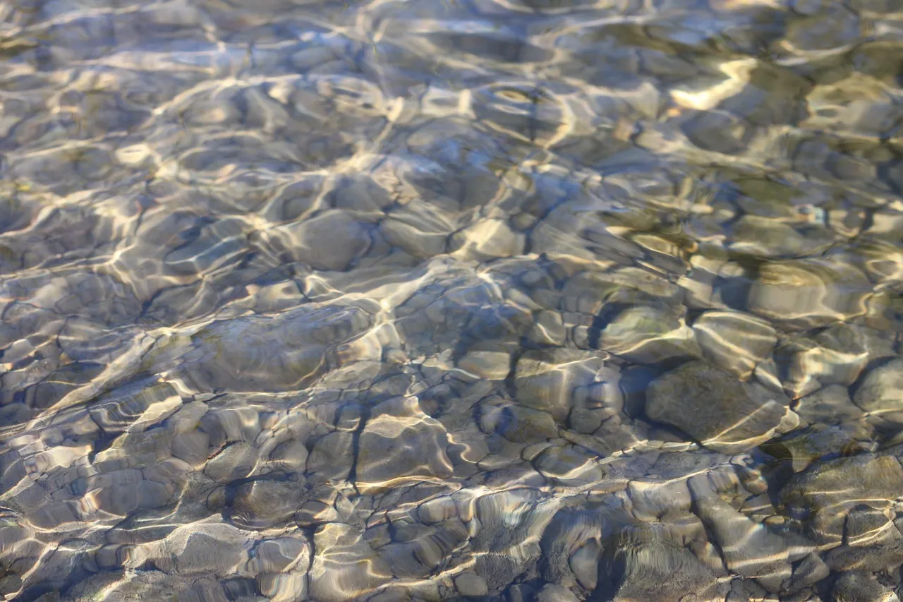 Sunlight rippling across clear shallow water over a bed of smooth grey stones