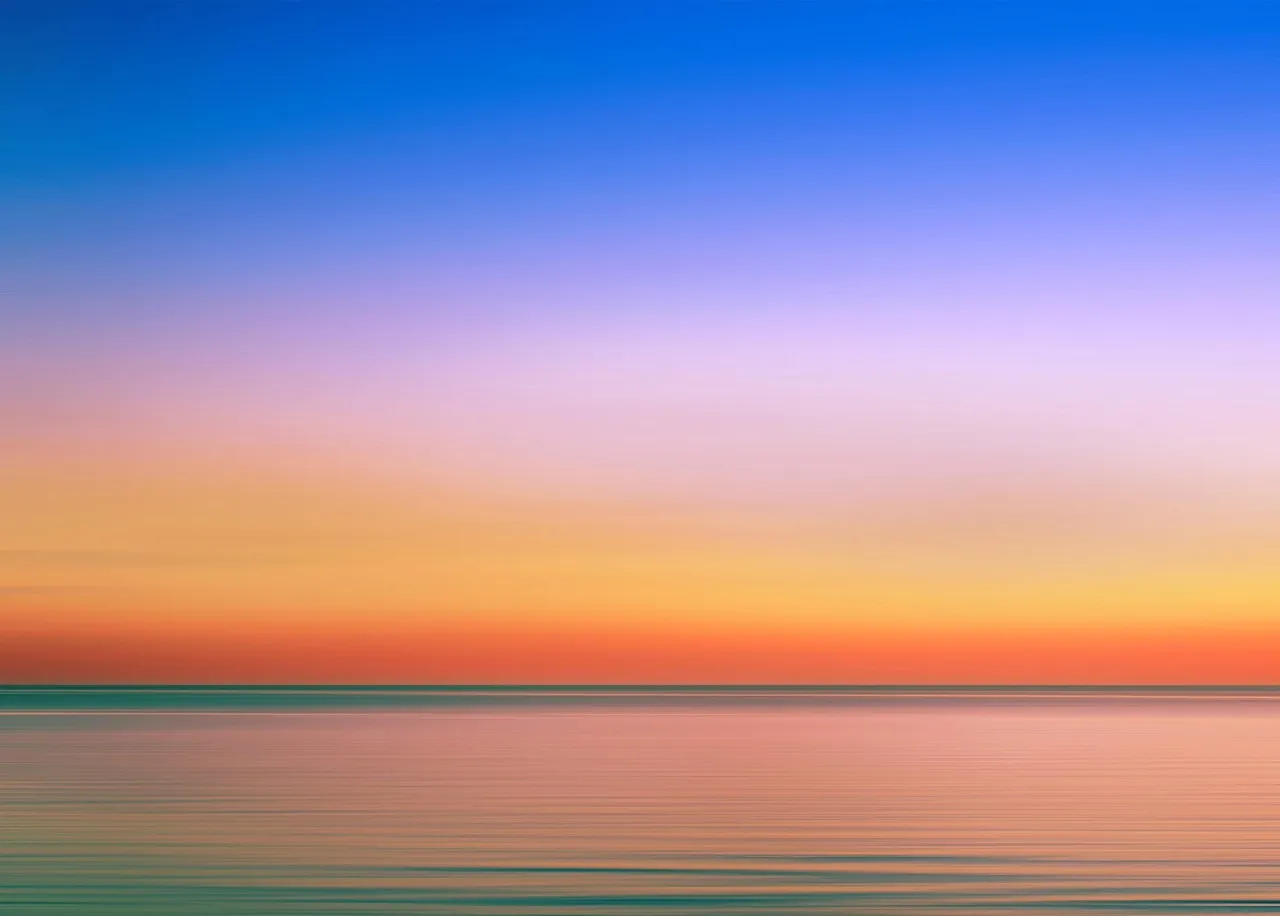 A long exposure of a flat, calm sea under a sky fading from blue through lilac to orange at the horizon