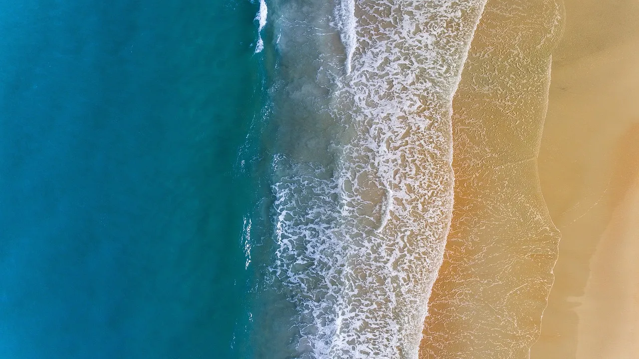 Looking straight down on a sandy shore, where turquoise water meets a band of white foam and golden sand