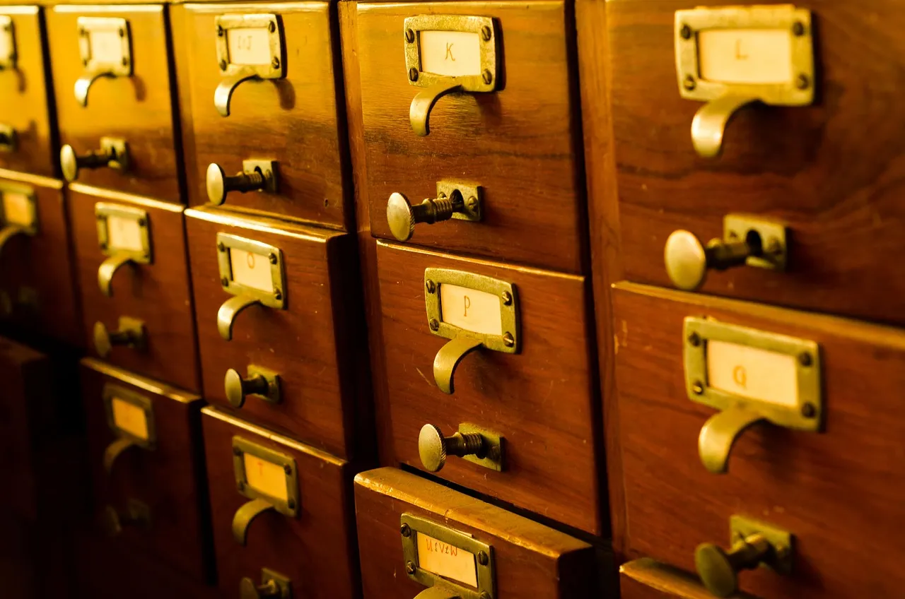 Rows of wooden catalogue drawers with brass handles and card labels marked K, L, P and Q, lit warm from one side.