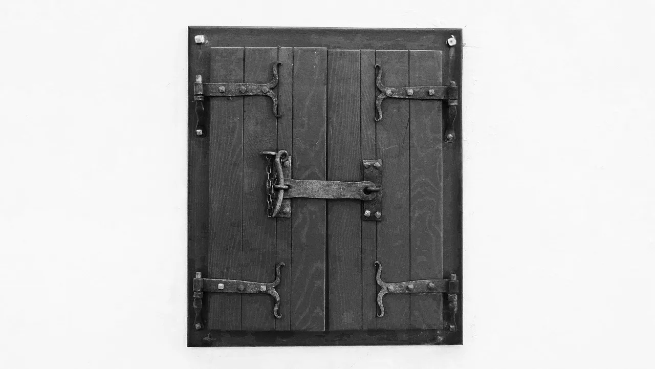A black and white photograph of a small pair of wooden shutters on a white wall, each leaf hung on two iron strap hinges, with one iron bar latching the two leaves together in the middle.