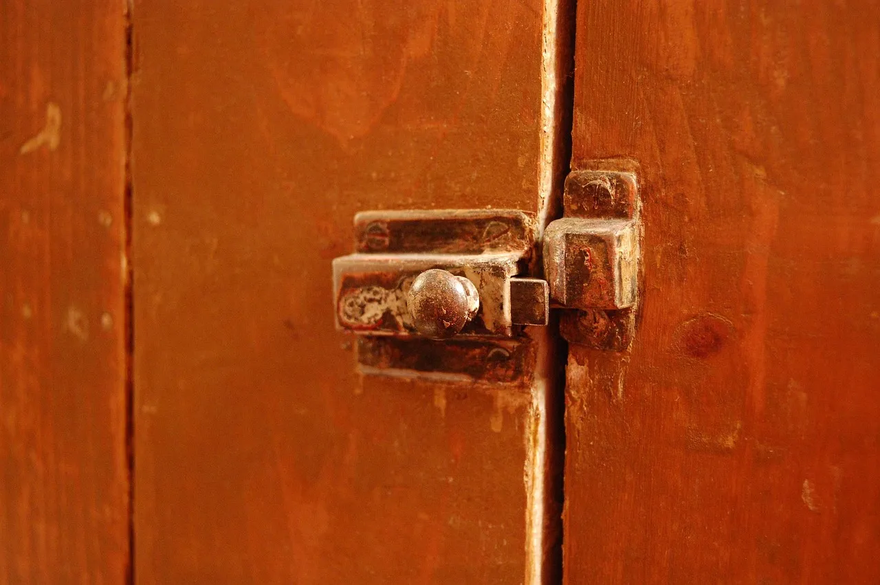 A close-up of an old metal slide bolt on a pair of orange-brown wooden doors, its bolt pushed across the gap into a keeper on the other door.