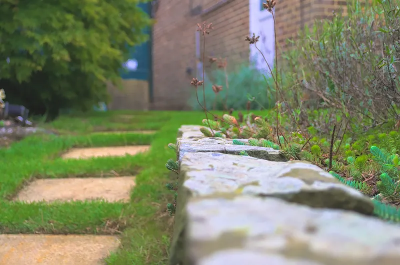 Stepping-stone pavers set in a lawn beside a low stone garden wall, with a brick building out of focus behind