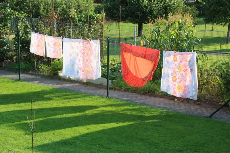 Sheets and pillowcases hanging on a washing line across a lawn, beside a paved path and a wire fence