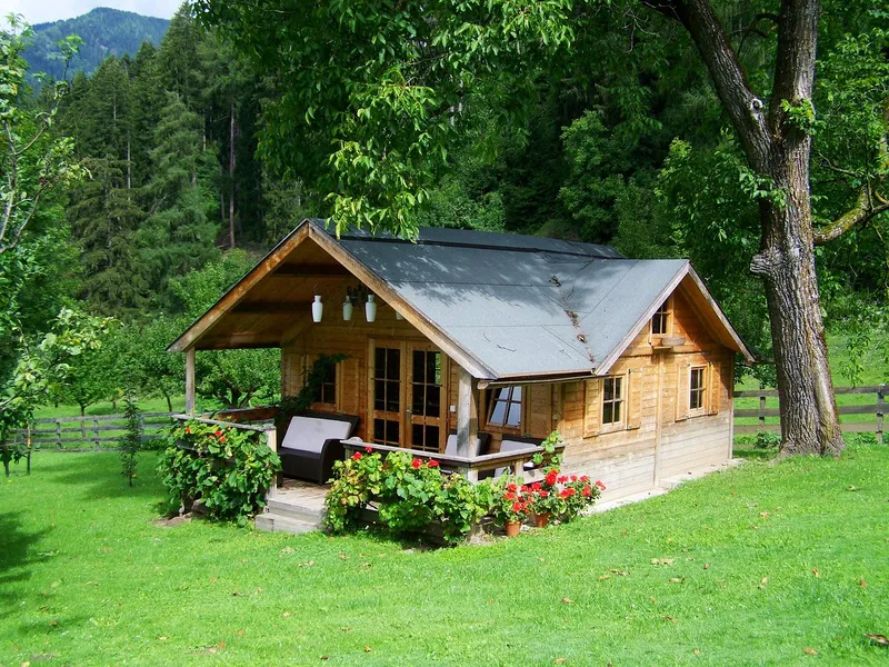 A small timber cabin with a grey roof and a covered porch with outdoor seating and red geraniums, on a lawn under a large tree