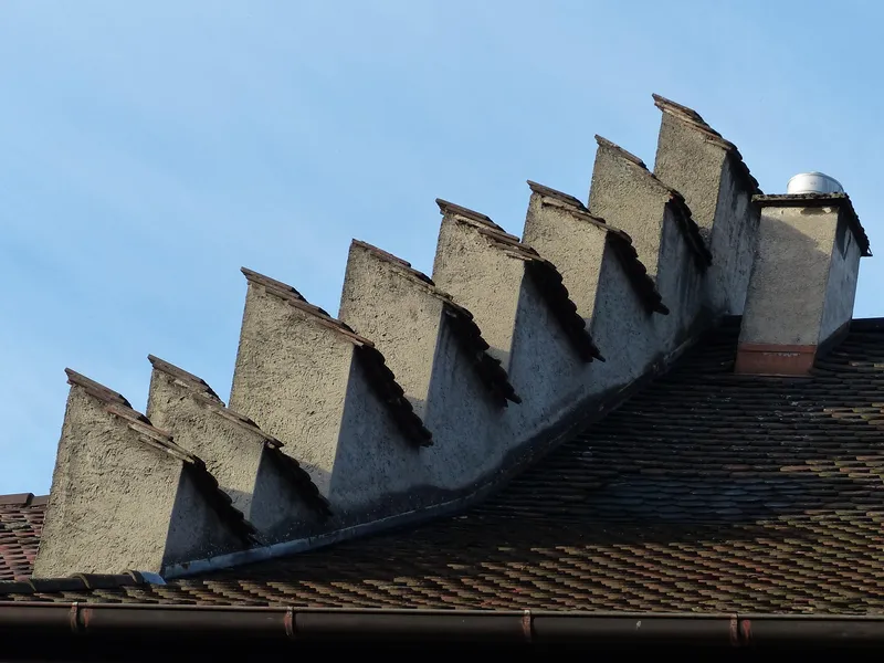 A stepped, rendered masonry wall rising above a tiled roof against a blue sky, with a chimney at its upper end.