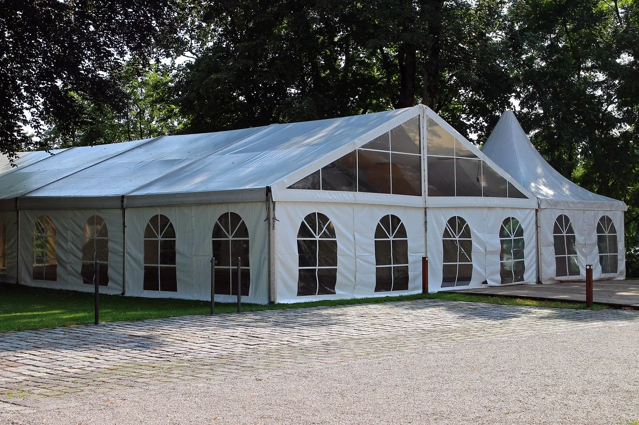 A long white marquee with arched clear windows, joined to a pointed pagoda tent, standing on grass beside a gravel forecourt under tall trees.
