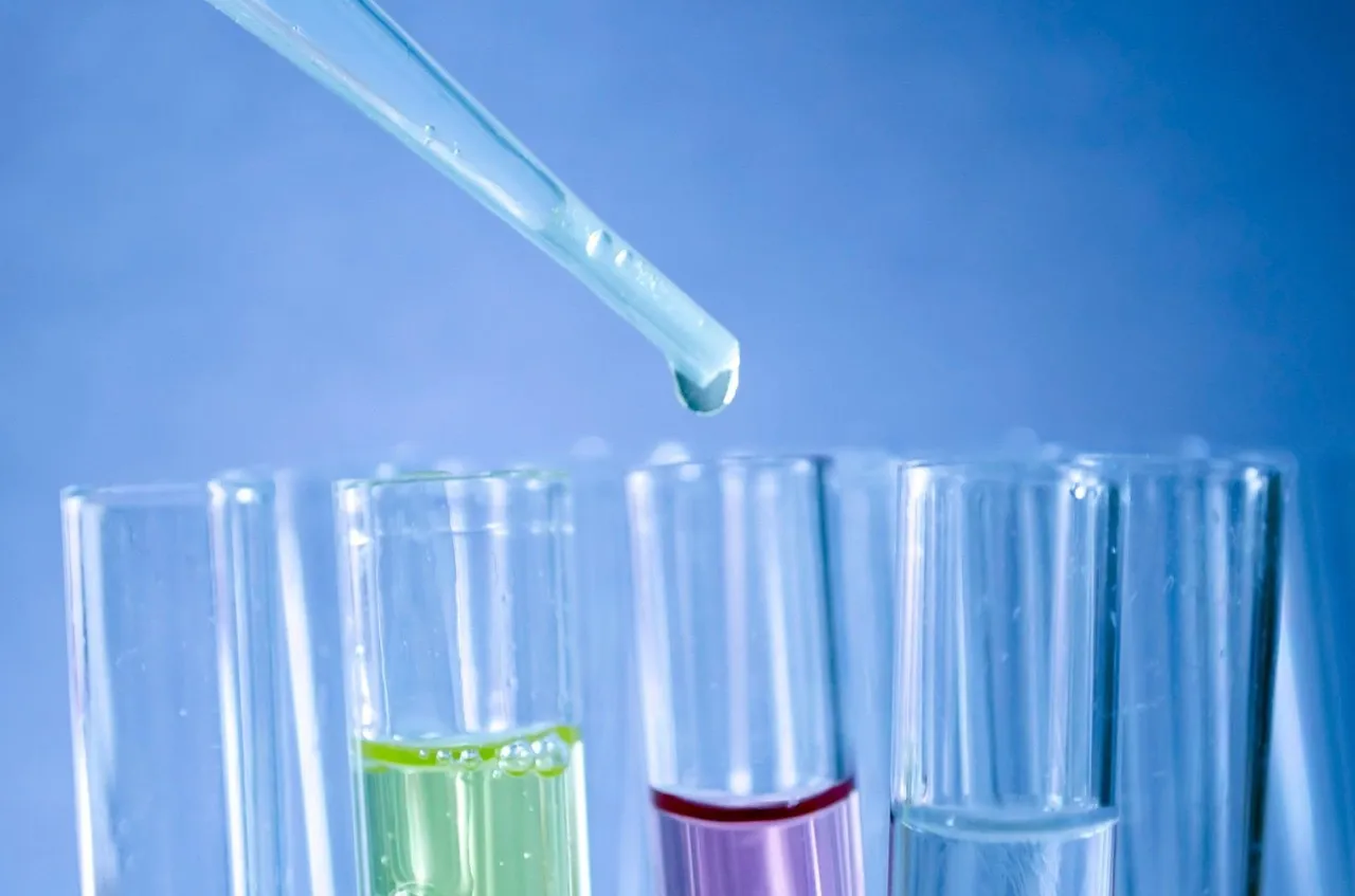 A glass pipette holding a drop above a row of clear test tubes, three of them part-filled with green, pink and clear liquid, against a pale blue ground