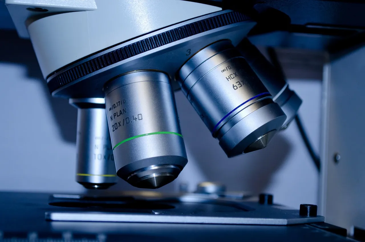 Close-up of a laboratory microscope’s objective lenses, marked 10x, 20x and 63x, above the stage, in cool blue light