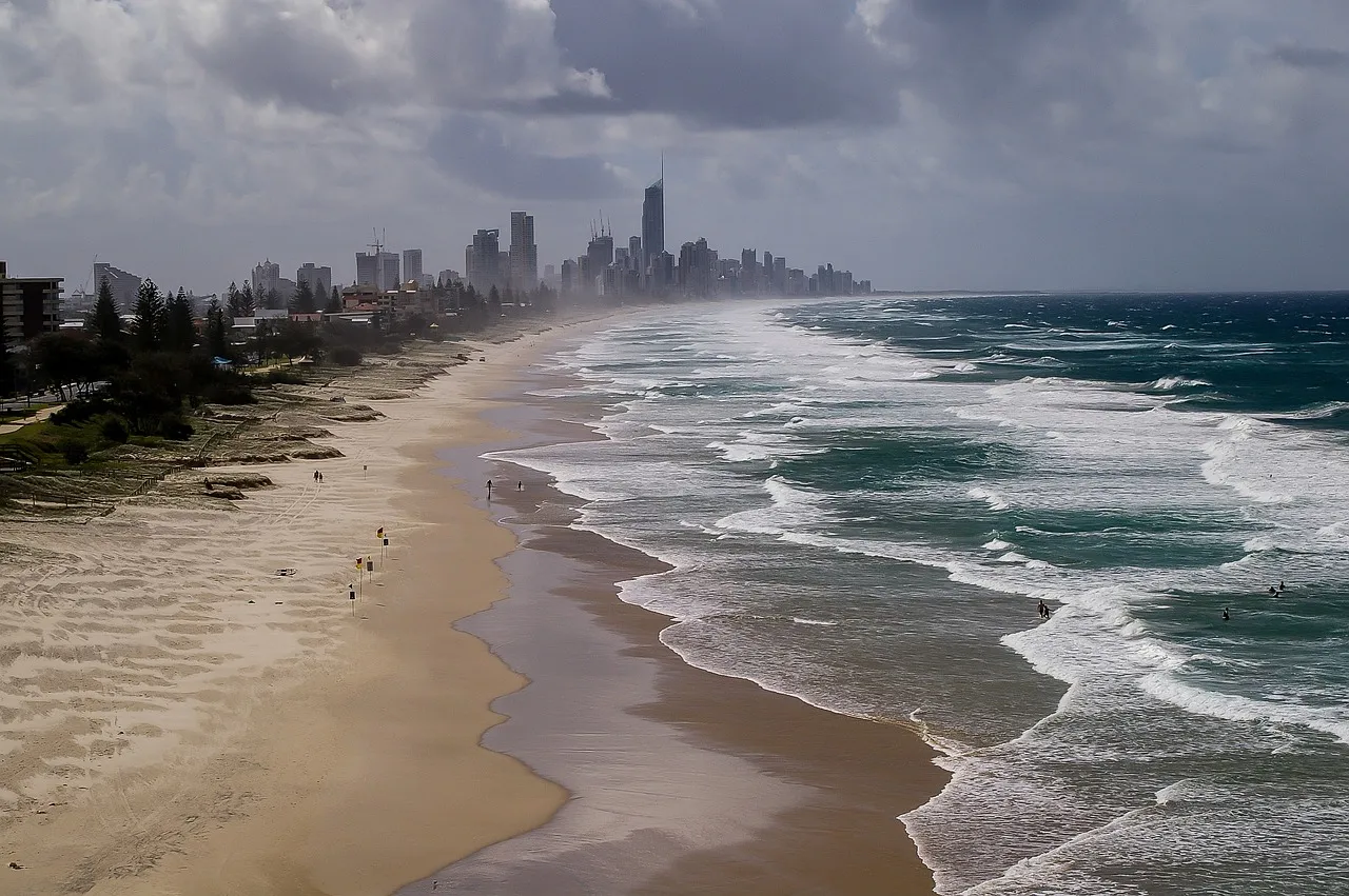 A long Gold Coast beach seen from above under heavy cloud, wet sand along the waterline, a few red and yellow flags on the sand, and high-rise towers in the distance
