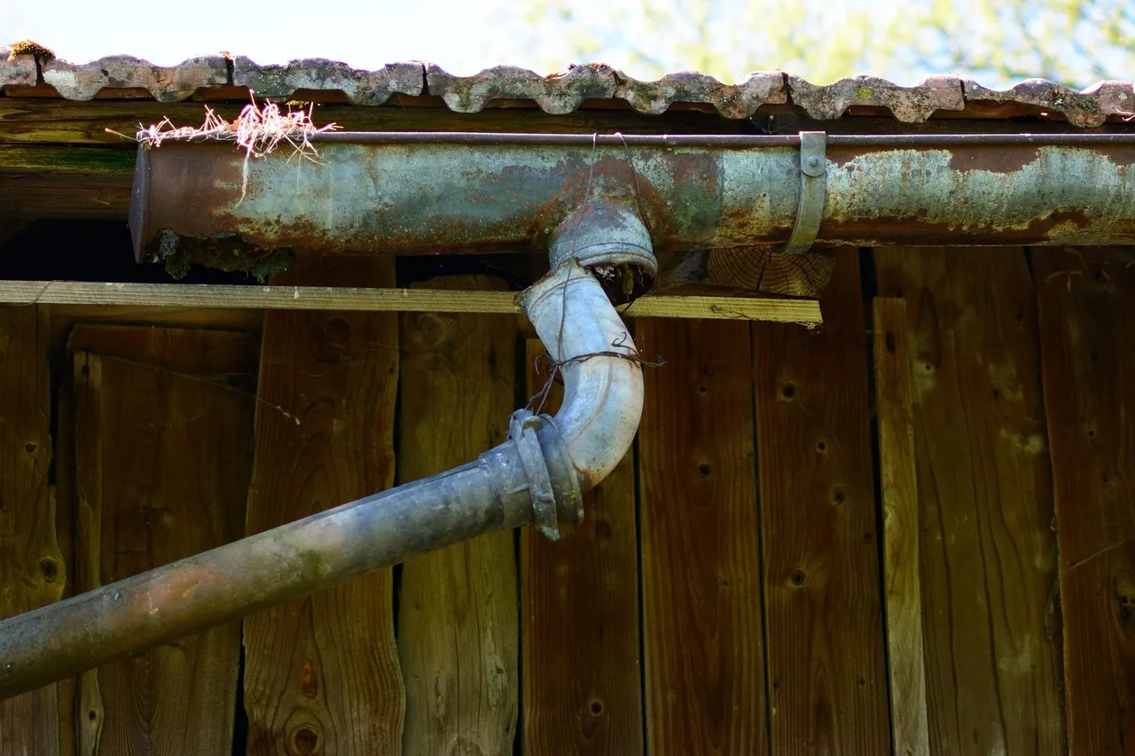 A weathered, rust-streaked metal gutter under a corrugated roof edge, its outlet joined to a downpipe elbow held together with twisted wire against old timber boards