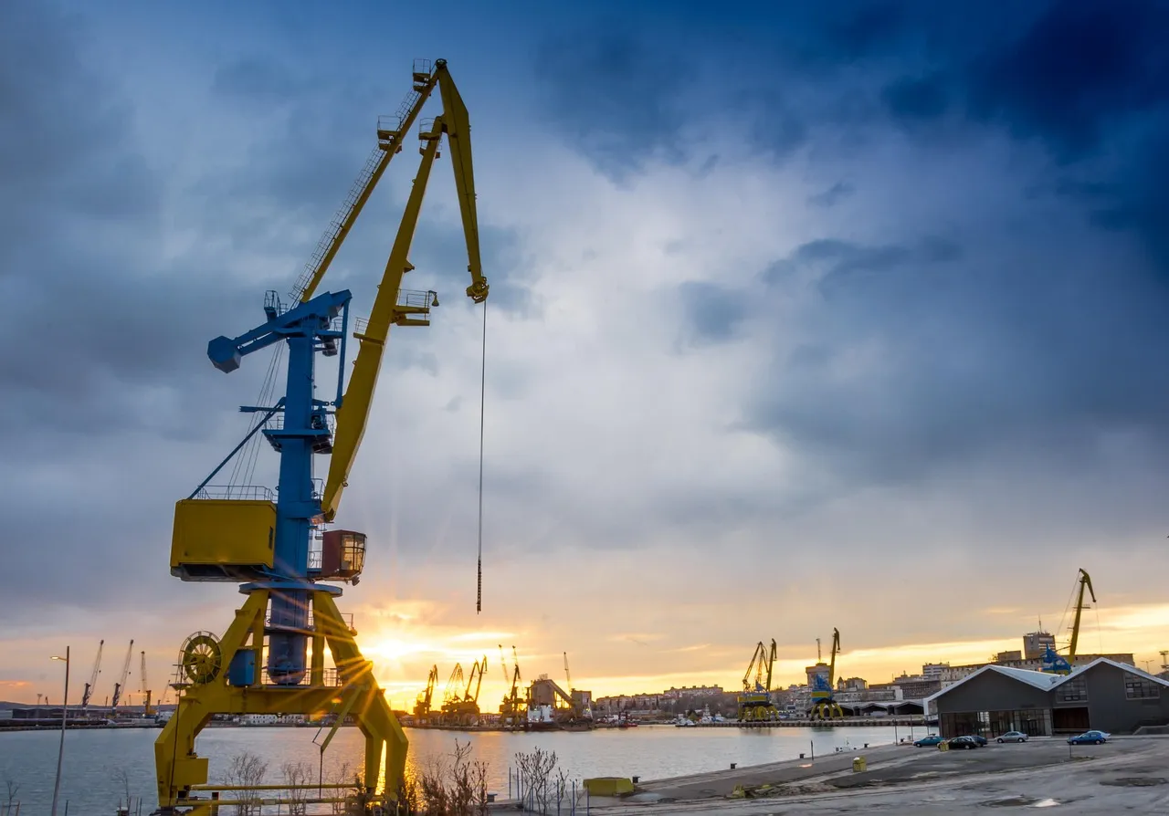 A tall harbour crane, painted yellow and blue, against a cloudy sky at sunset, with a quay and more cranes along the far shore.