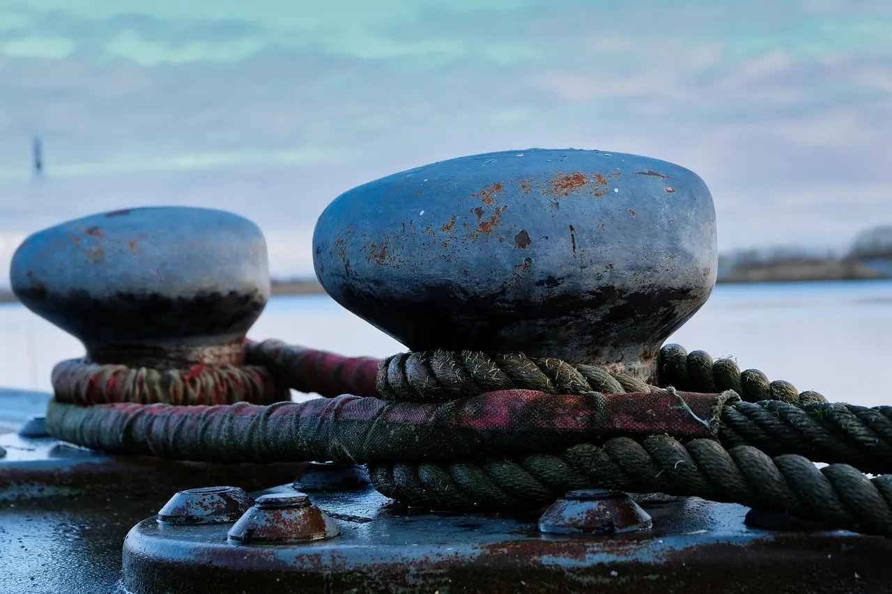 Two weathered iron mooring bollards on a quay, wound with thick ropes, with water and a pale sky behind.