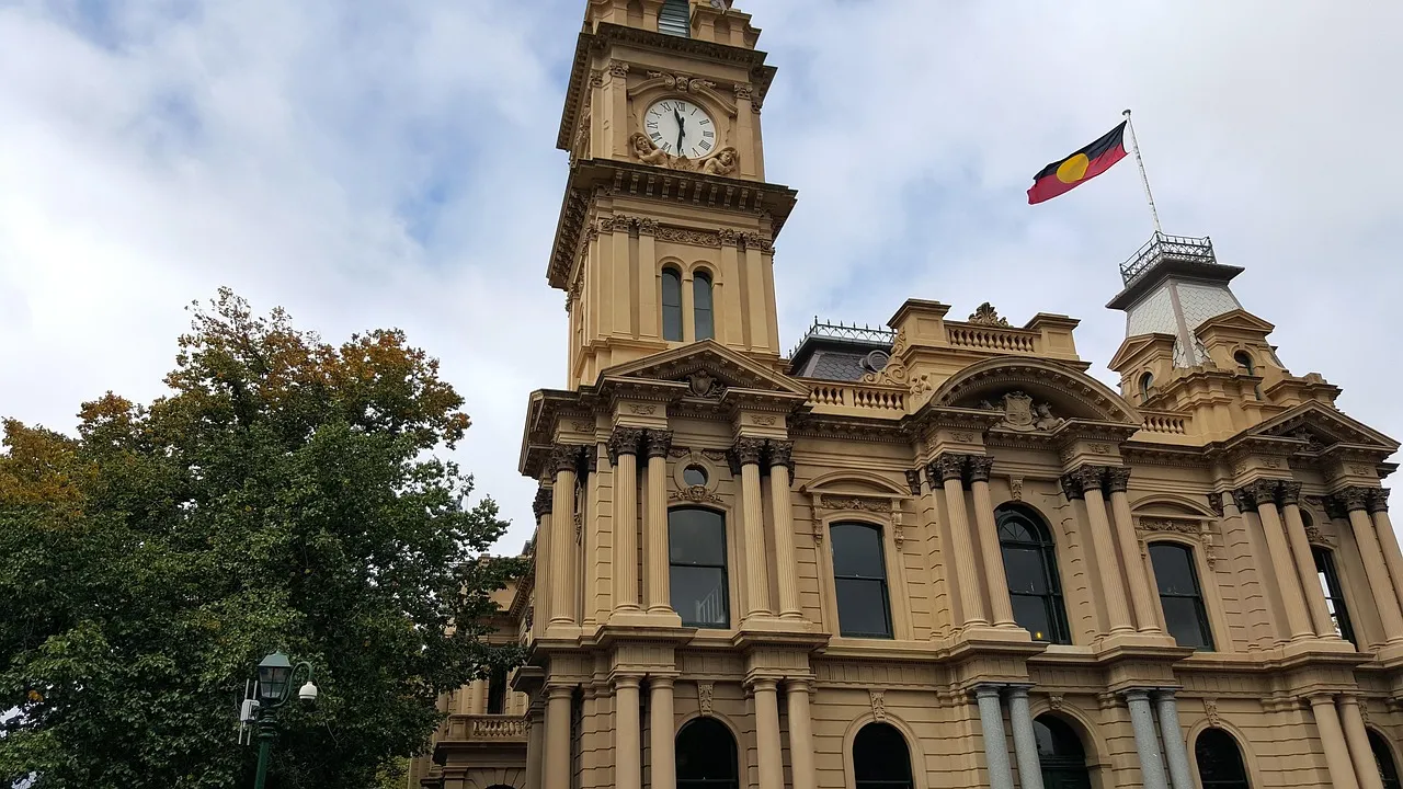 The clock tower and sandstone-coloured facade of Bendigo Town Hall under a cloudy sky, with the Aboriginal flag flying from the roof