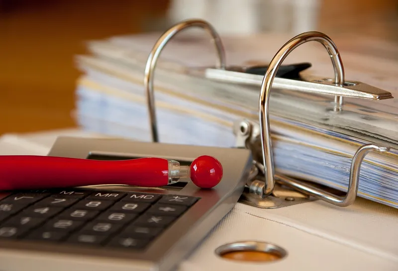 An open lever-arch file packed with papers, beside a calculator with a red pen lying across it.