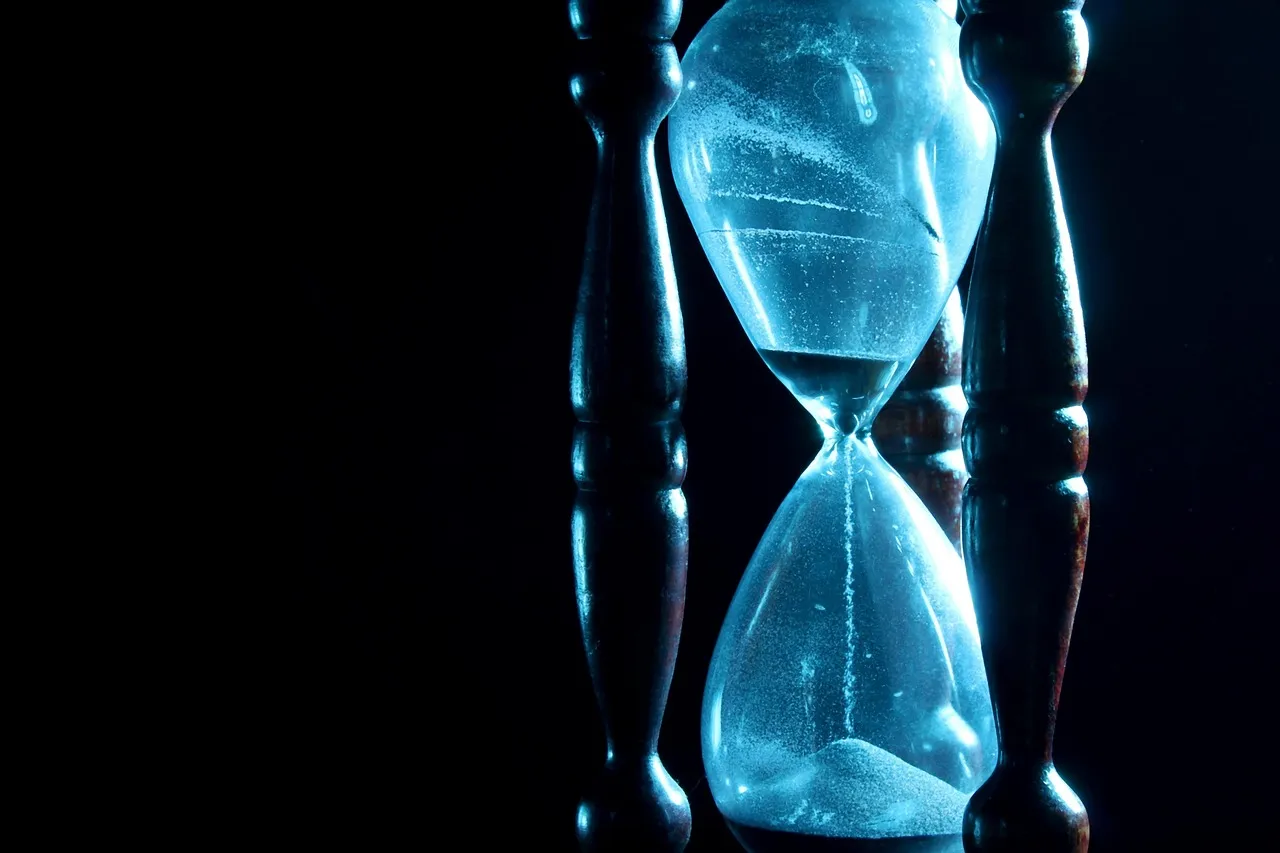 A wooden-framed hourglass lit in cold blue against a black background, sand running into the lower bulb.