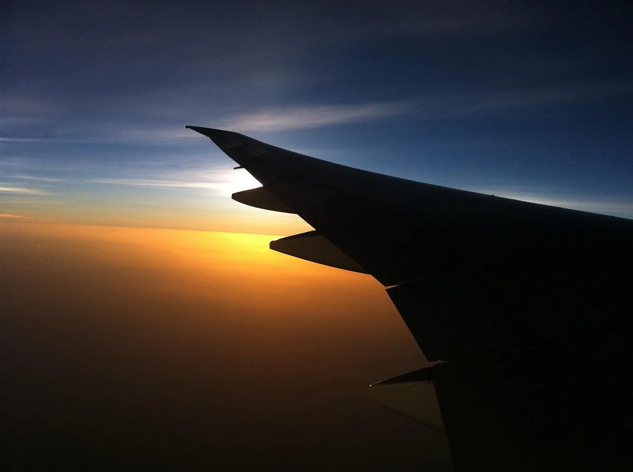 An aircraft wing in silhouette against an orange sunrise, seen from a passenger window above the clouds