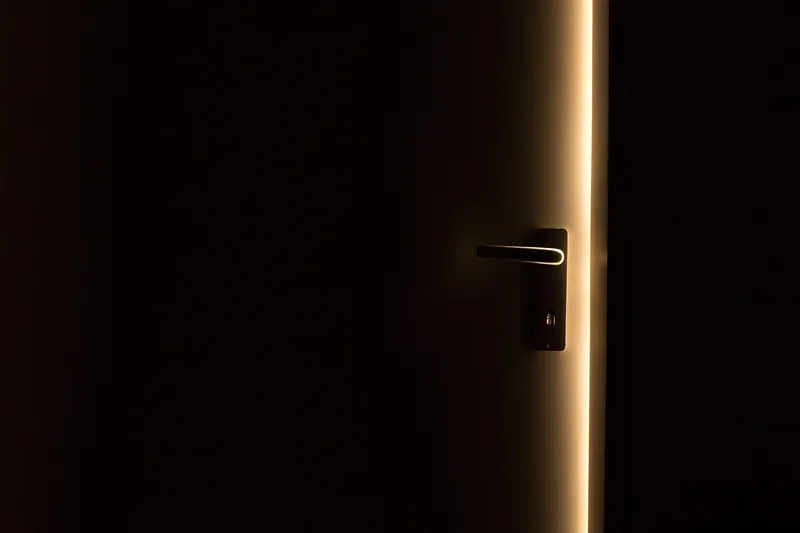 A dark door standing slightly open, a thin line of warm light along its edge and across the handle.