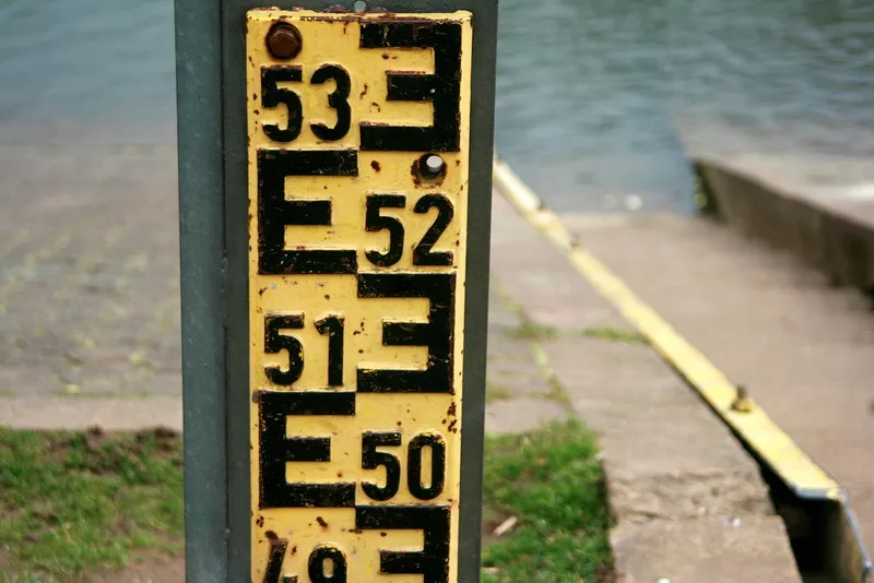 A yellow river gauge board with black E-shaped marks and painted numbers from 49 to 53, fixed to a post beside a boat ramp running down into grey water.
