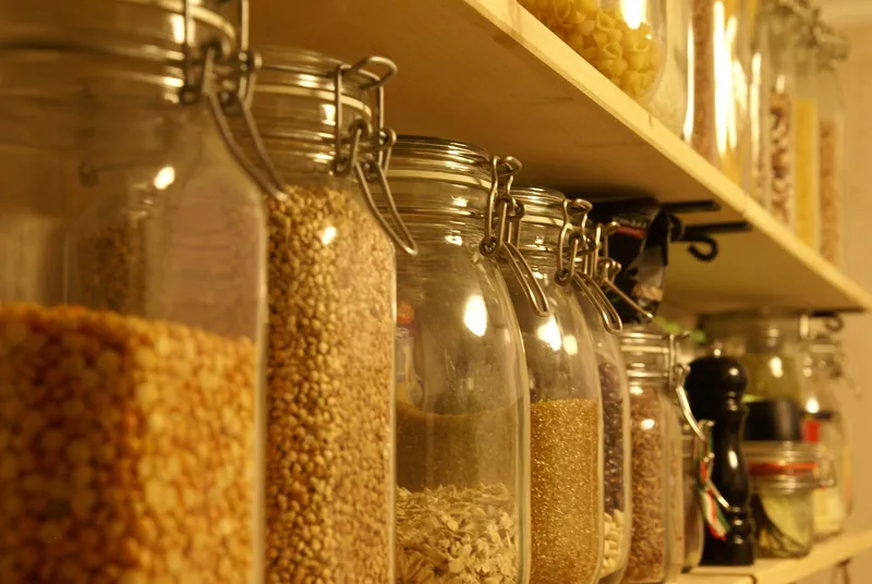 Glass jars with clip-down lids lined up on a wooden pantry shelf, filled with dried peas, grains and oats, with pasta in jars on the shelf above.
