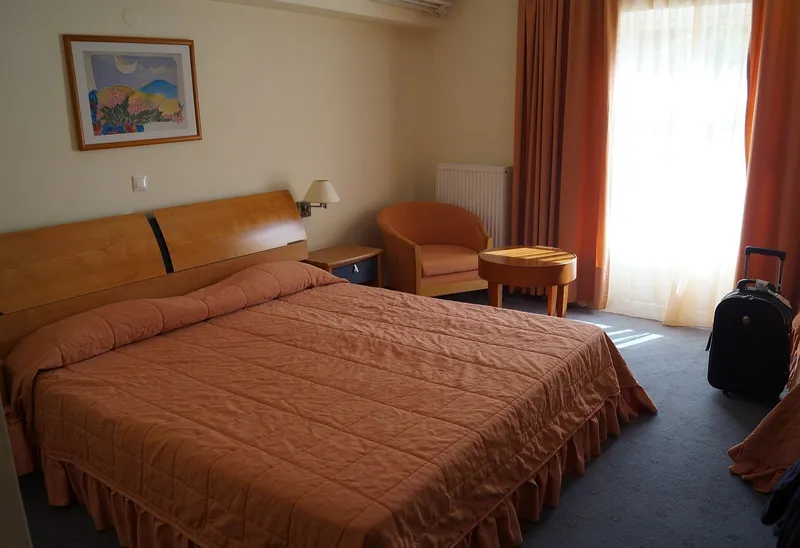 A hotel room with a double bed under an orange quilted cover; a black wheeled suitcase stands on the carpet by the window, away from the bed.