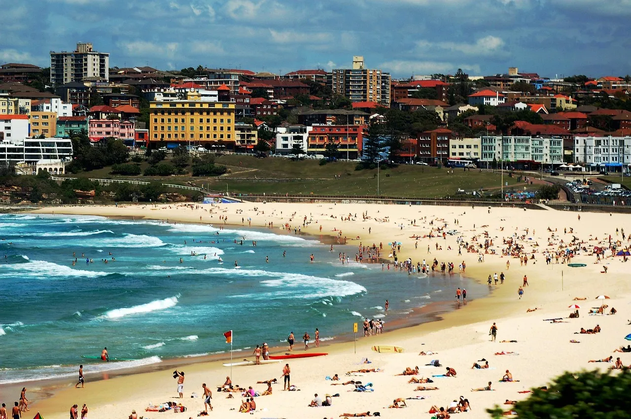 A crowded surf beach with a red and yellow flag on the sand, and apartment blocks and red-roofed buildings packed on the slope behind it