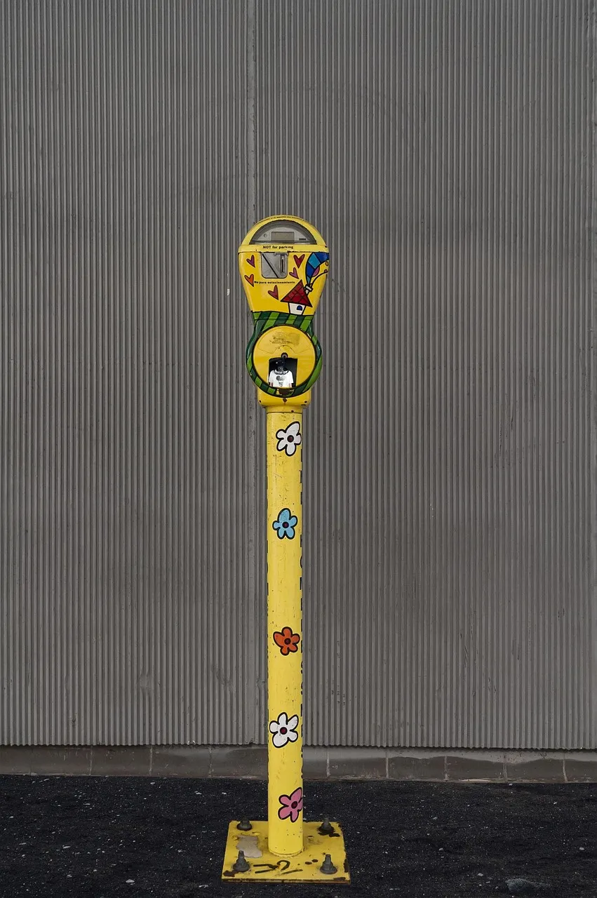 A parking meter on a yellow post painted with flowers and a little house, standing against a grey corrugated wall