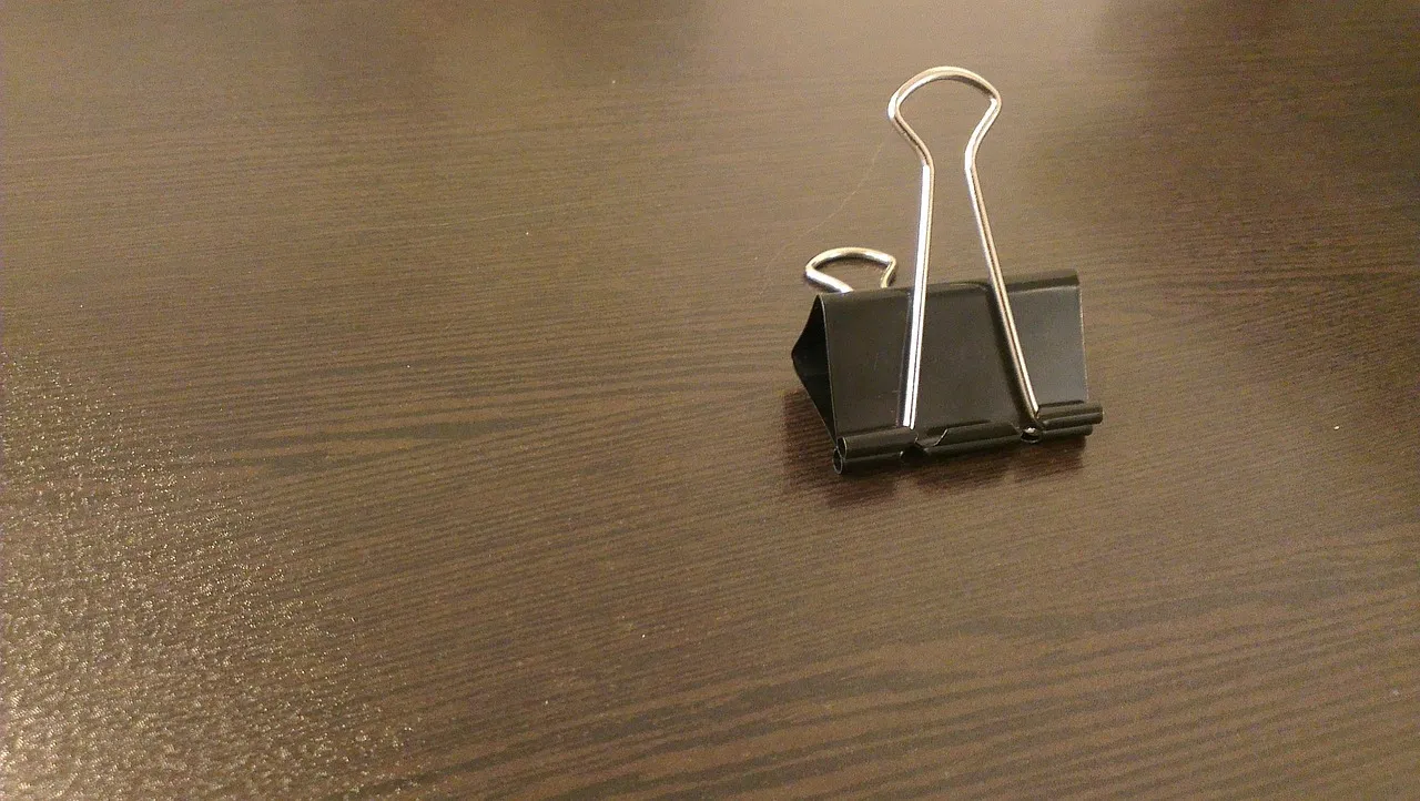 A black binder clip standing open on a dark wooden desk.