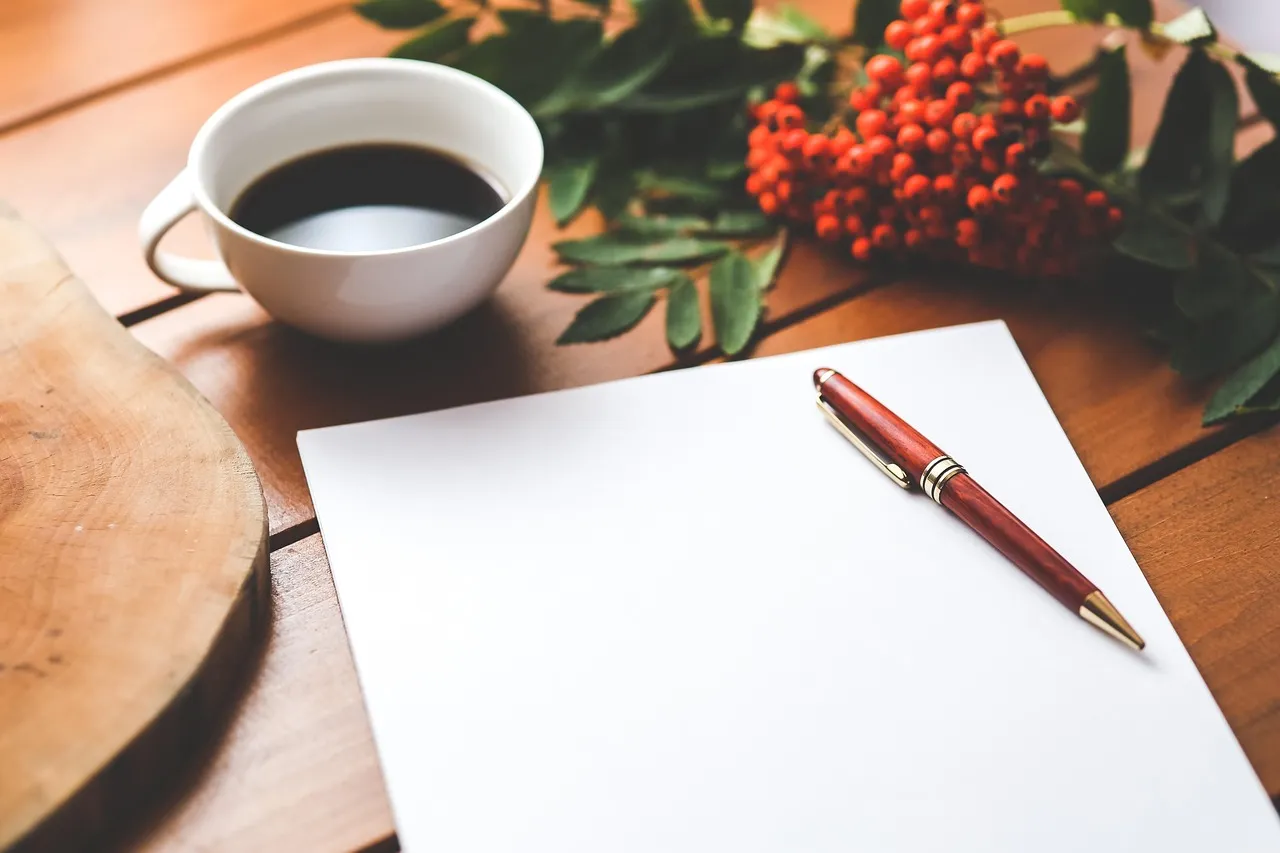 A blank sheet of paper and a wooden pen on a table, beside a cup of black coffee and a sprig of red berries.