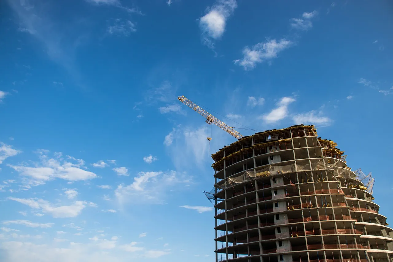 A rounded concrete building under construction, its upper floors open to a blue sky, with the jib of a tower crane above it