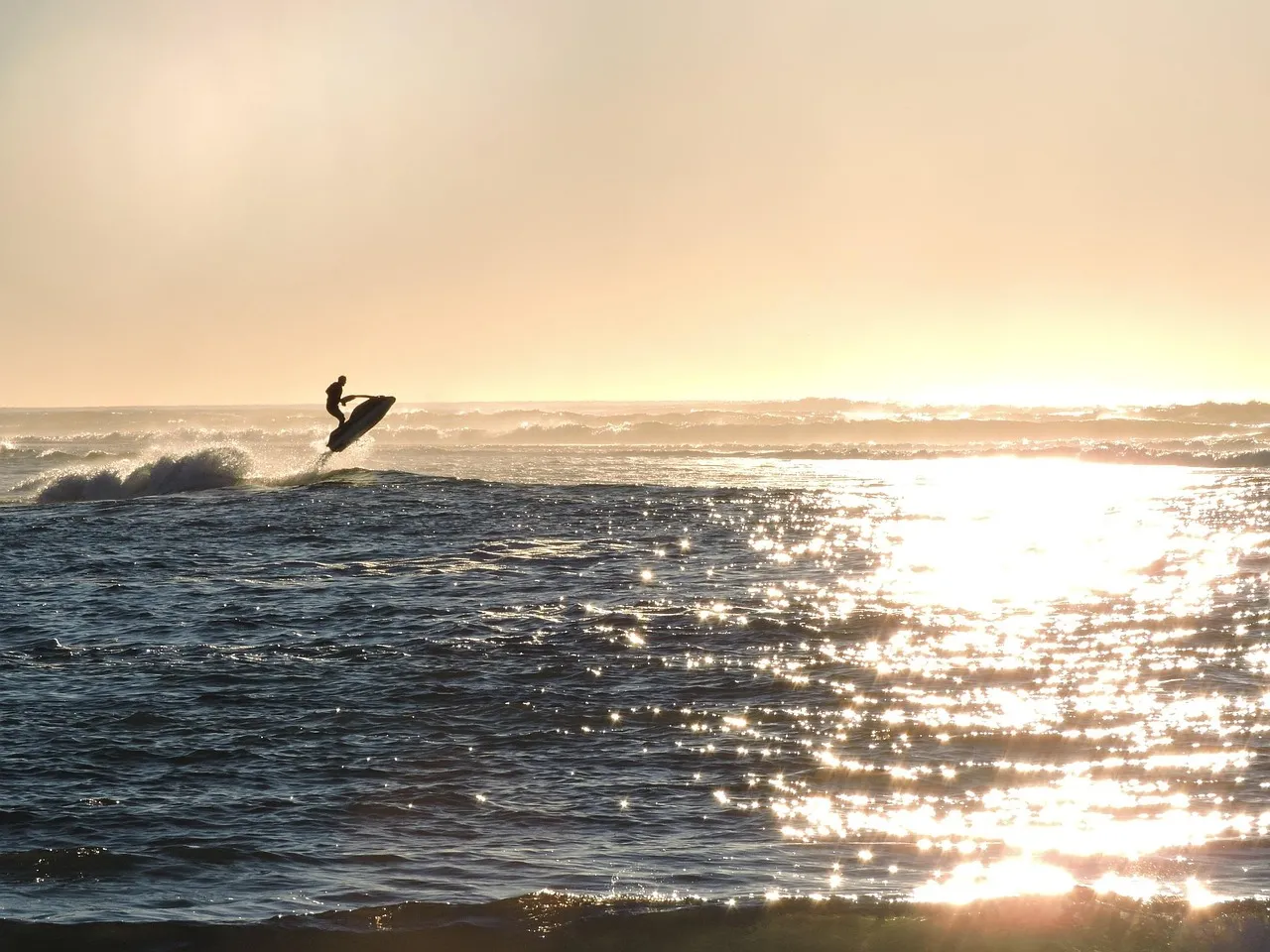 A rider on a jet ski in silhouette, lifted off a breaking wave, with the low sun glittering across the sea.