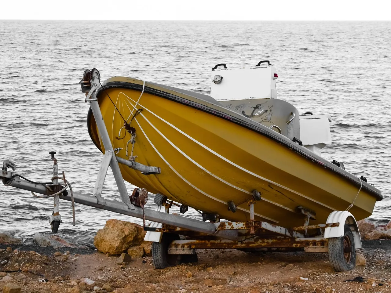 A small yellow open boat with a white console, resting on a weathered boat trailer on stony ground at the water’s edge, with grey sea behind. Everything but the boat is in black and white.