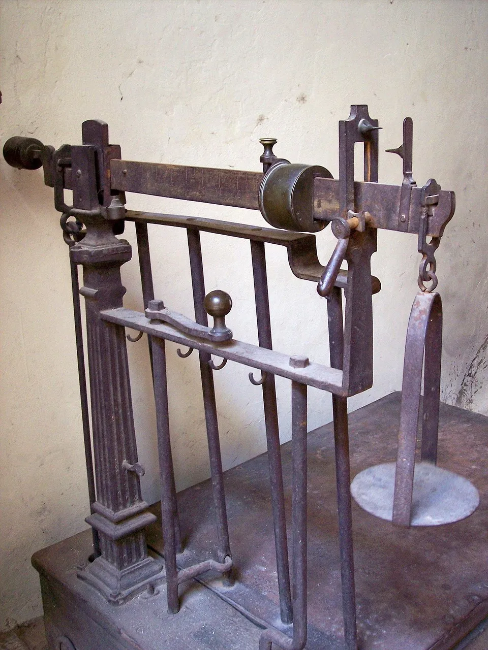 An old iron beam scale against a pale wall. A brass weight sits on its graduated arm, a hanger with a round plate hangs from one end, and the frame stands on a low iron platform.
