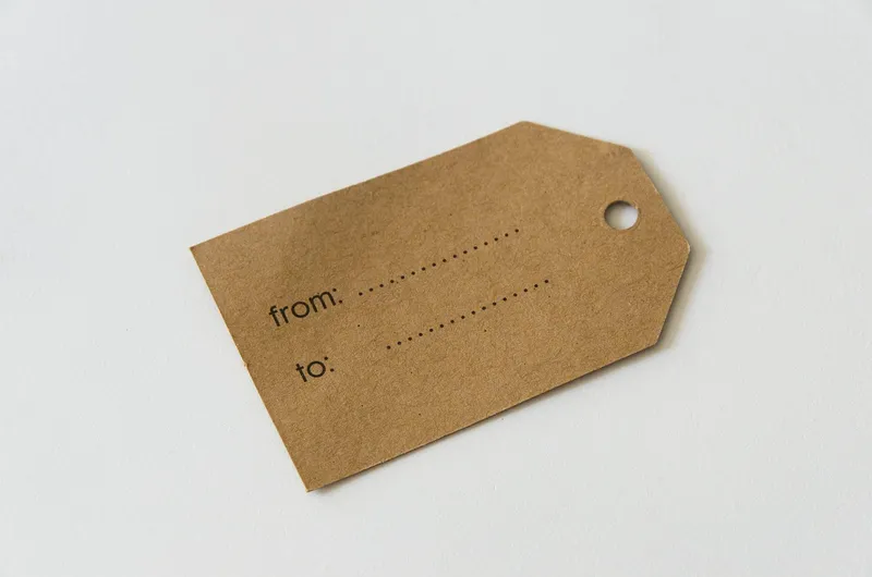 A blank brown paper tag with a punched hole, printed with empty from and to lines, lying on a white surface