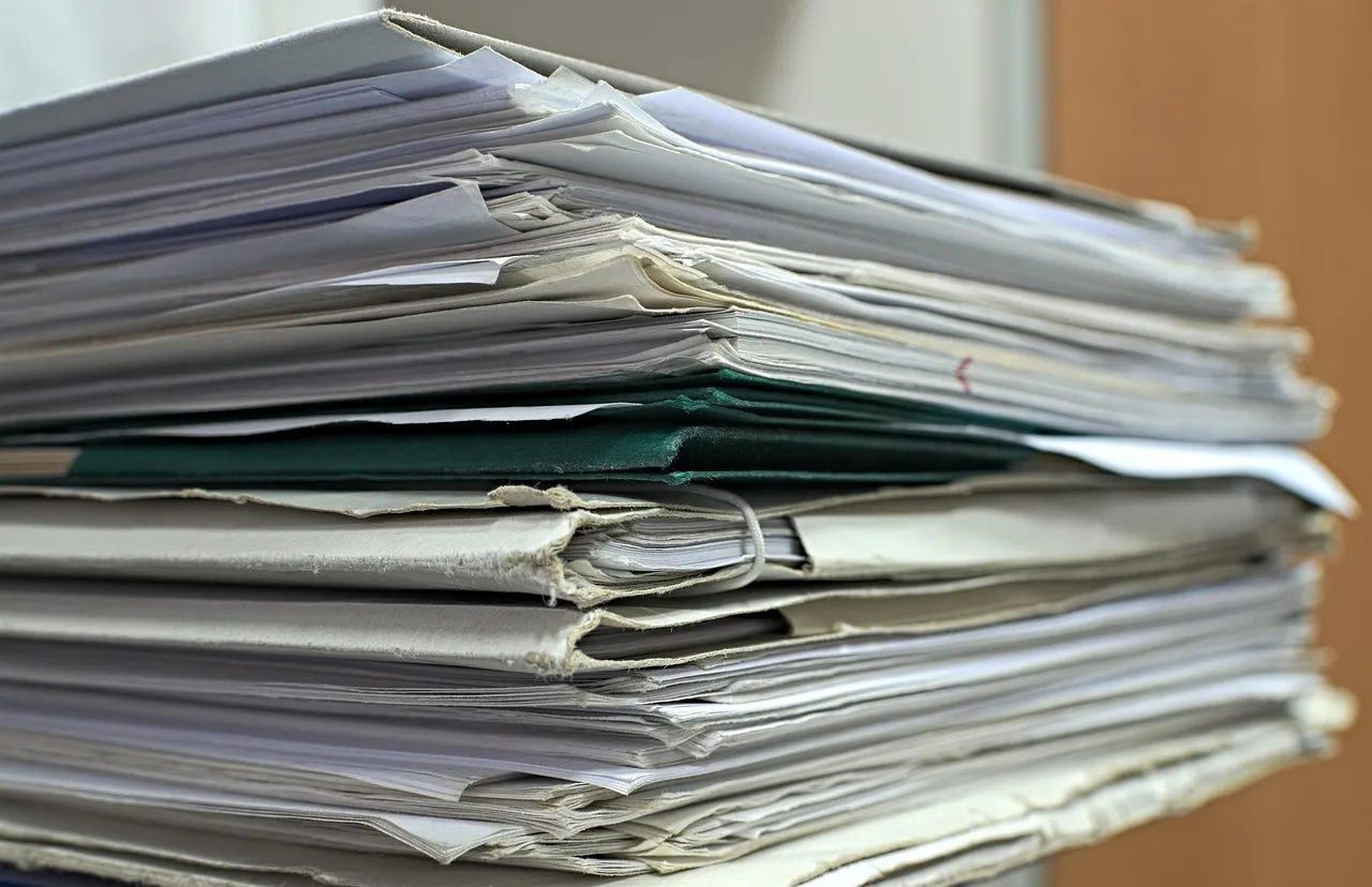 A tall stack of paper files and folders, one dark green folder among them, on a desk