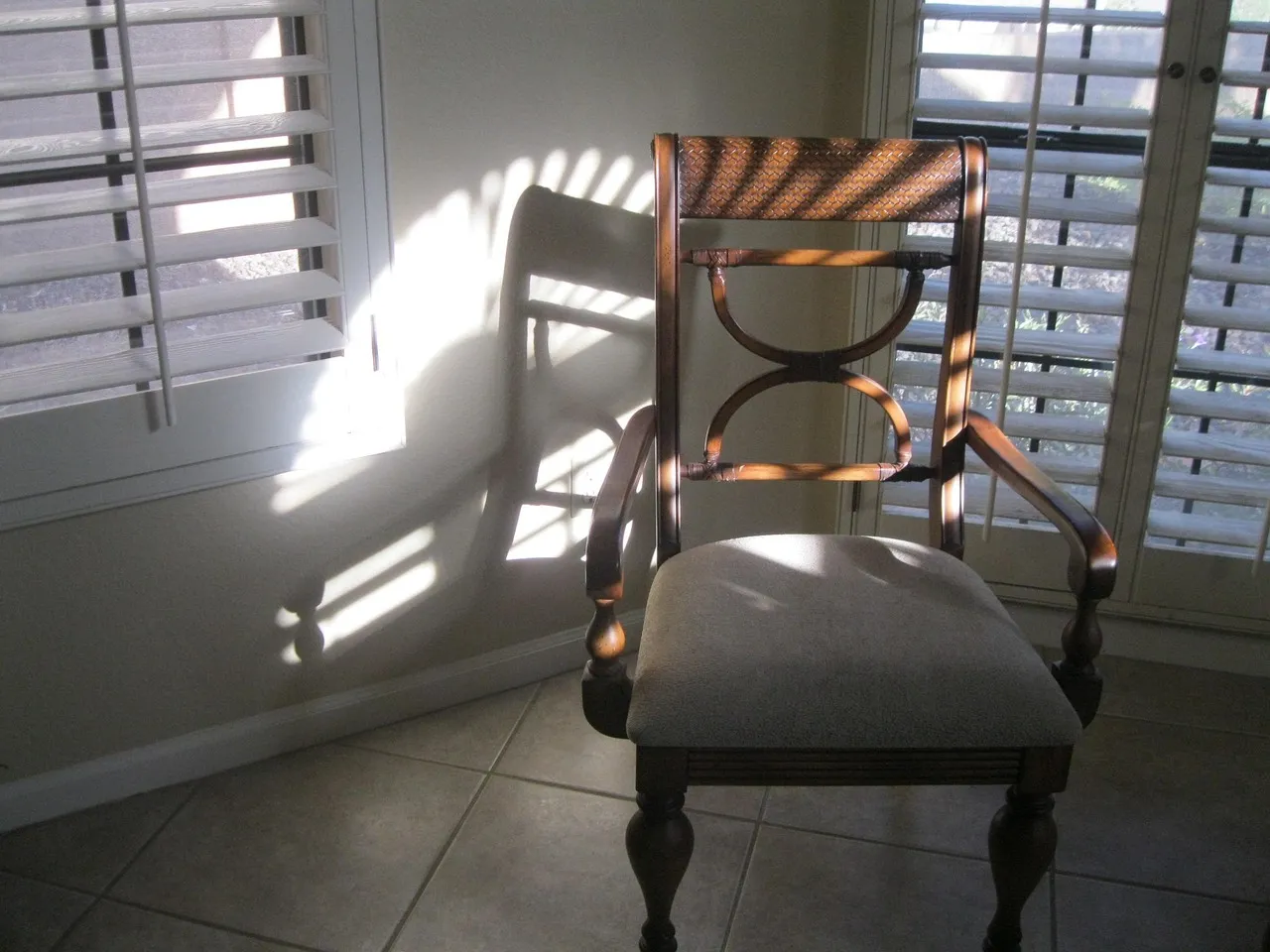 Late sun through white louvred shutters throws bands of light and the shadow of a wooden chair across a tiled room.