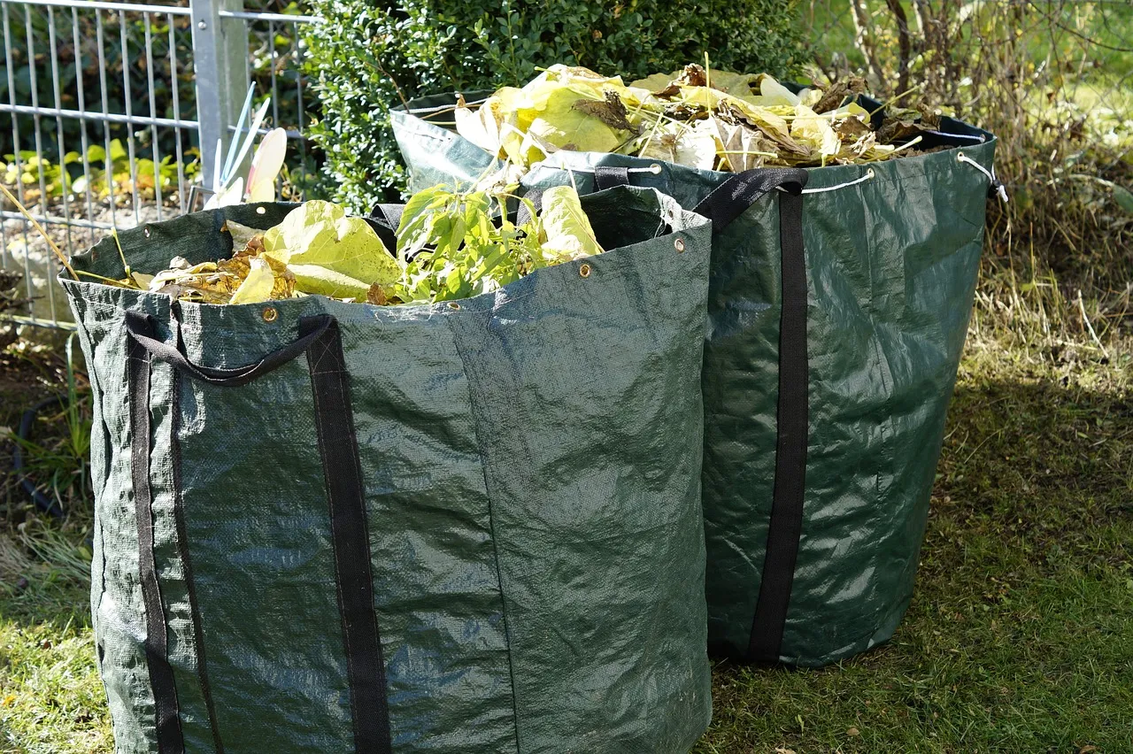 Two green woven garden bags on a lawn, filled to the top with leaves and plant cuttings.