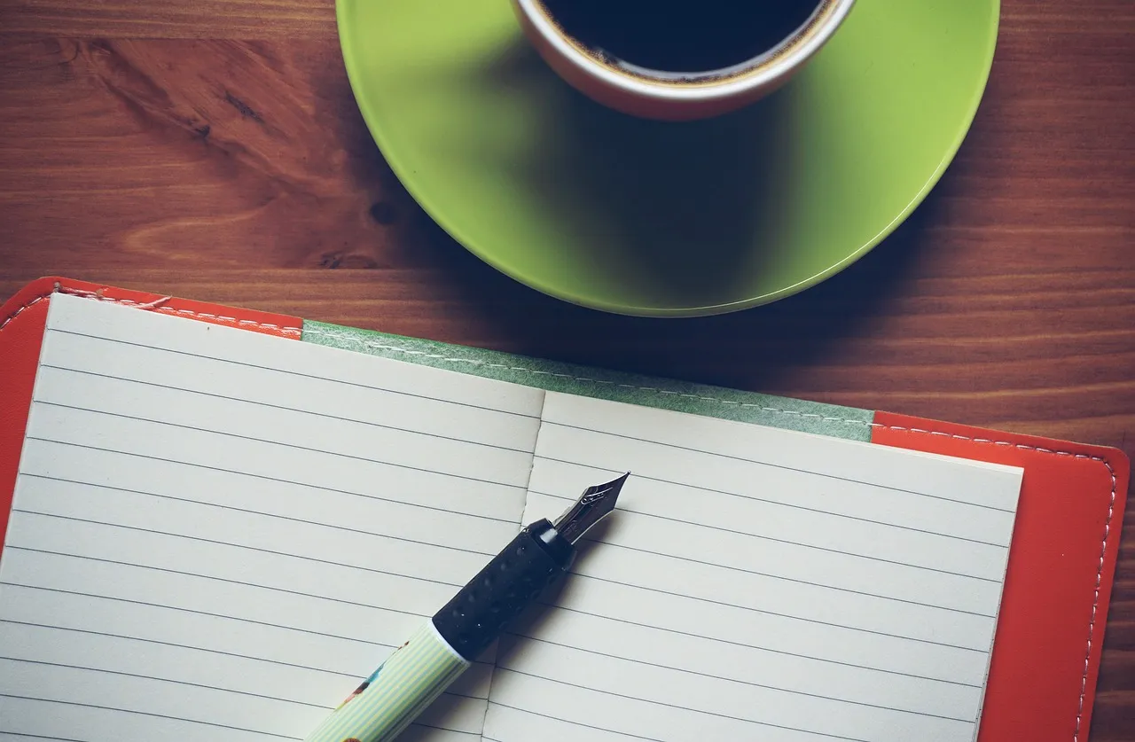 An open notebook in a red cover, its ruled pages blank, on a wooden table, with a fountain pen resting on it and a cup of black coffee on a green saucer.