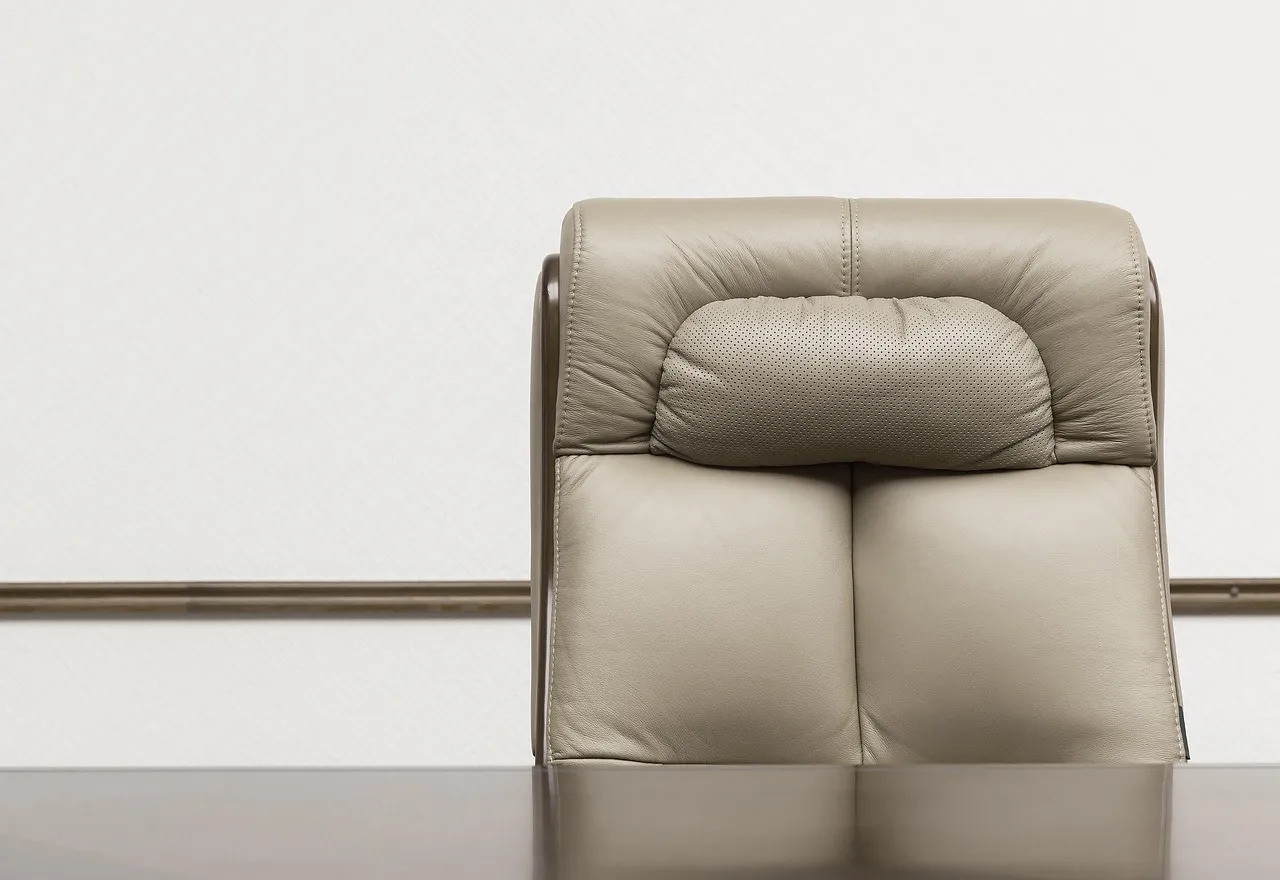 An empty beige leather chair behind a dark, polished table, against a pale wall with a wooden rail.