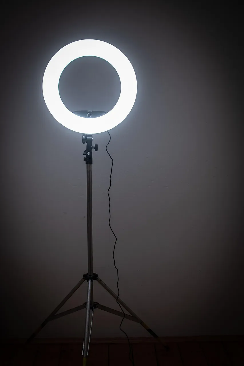 A lit ring light on a tall tripod stand in a dark, empty room, its cable trailing to the floor.